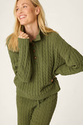 A smiling person with long blonde hair wears the P.J. Salvage Cozy Chenille Cable Knit Olivine Pullover, posing with one hand near their chin against a light background.