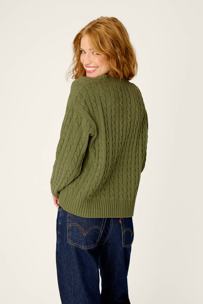 A woman with wavy red hair smiles over her shoulder, wearing the P.J. Salvage Cozy Chenille Cable Knit Olivine V-Neck Sweater and dark blue jeans against a plain light background.
