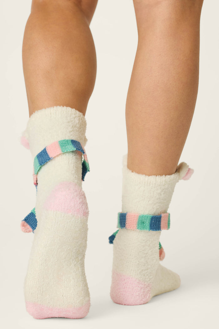 A person wearing P.J. Salvage Cozy Polar Bear Socks—fuzzy, cream-colored socks featuring pink toes and heels and pastel striped bands around the ankles—stands with their back to the camera.