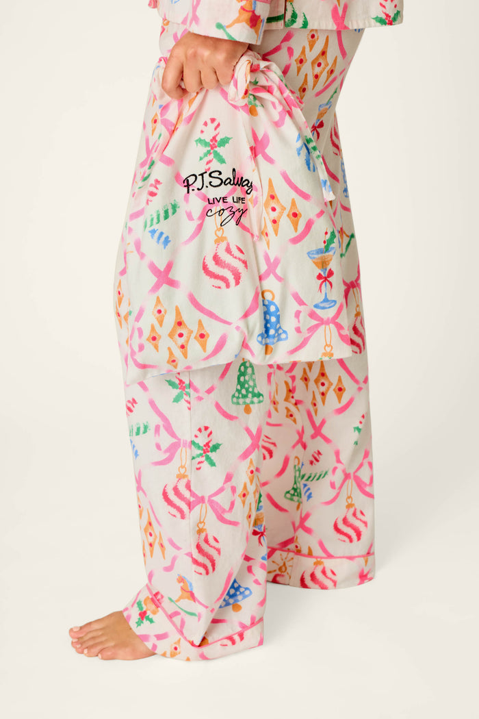 A person is wearing the Deck the Halls Ornament Print 4-Piece PJ Set by P.J. Salvage and holding a matching drawstring bag with the P.J. Salvage logo; their bare foot is visible against a plain white background.