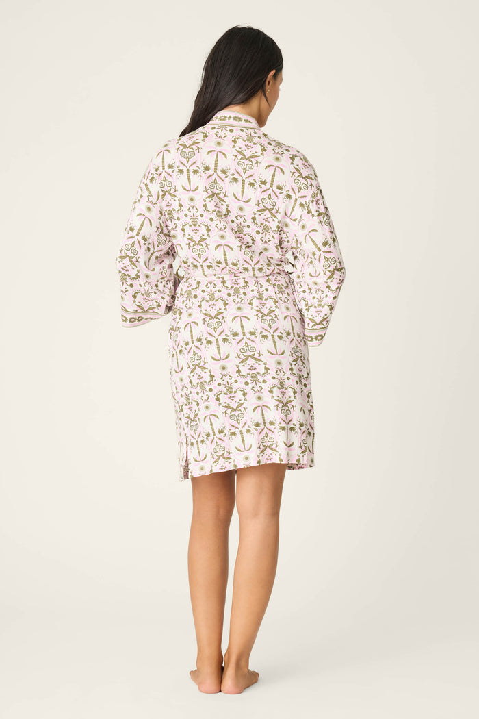 A woman stands barefoot facing away, wearing the Déjà Vu Dreams Tiki Time Robe by P.J. Salvage—a white robe with a pink and green floral pattern—against a plain light background.