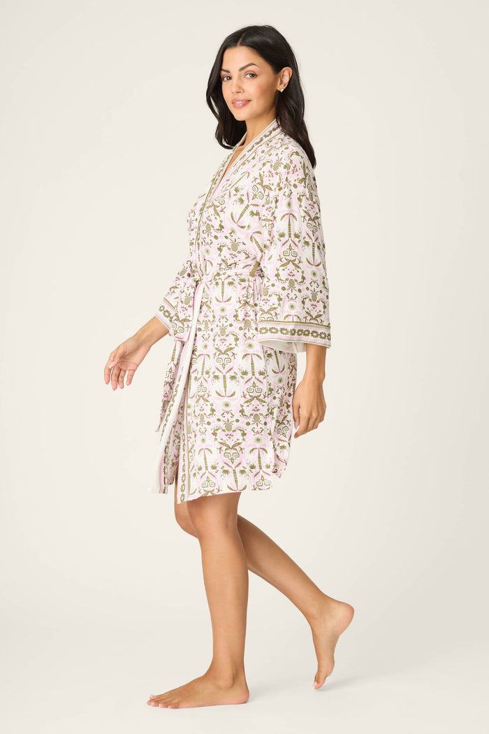 A woman with long dark hair wears the P.J. Salvage Déjà Vu Dreams Tiki Time Robe, a light floral robe with a belt and trim, standing barefoot, smiling and facing sideways against a plain background.