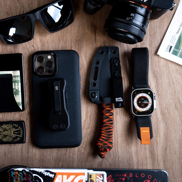 Discreet Carry Pocket Clip on a table with a phone, knife, watch, sunglasses, and camera, showcasing everyday carry essentials.