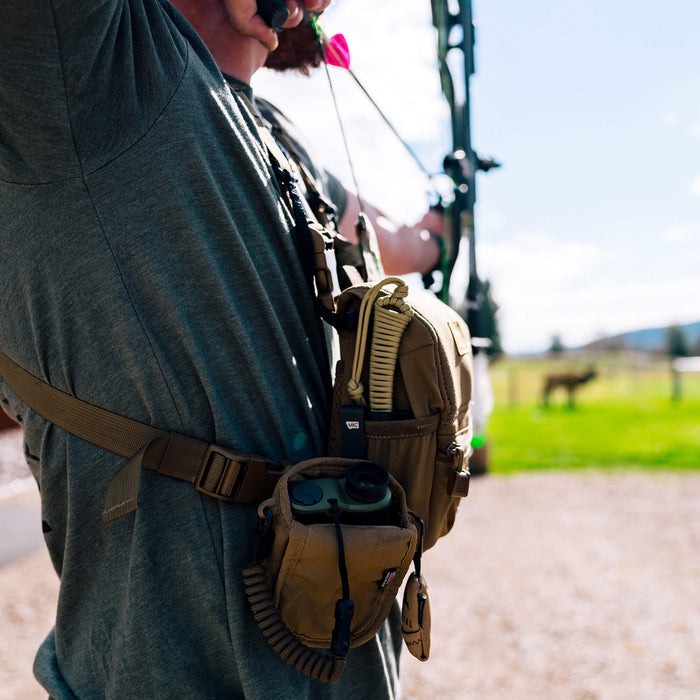 A person wearing a gray shirt uses a discreet carry pocket clip on a tan pouch while aiming a bow outdoors.