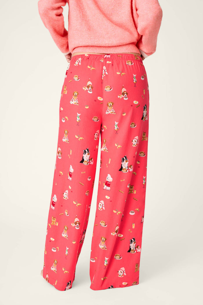 A person is shown from the waist down and viewed from the back, wearing P.J. Salvage Dogs Who Brunch Flannel Pajama Pants featuring a bright red background with a playful pattern of cartoon dogs and brunch foods, paired with a matching pink sweater.