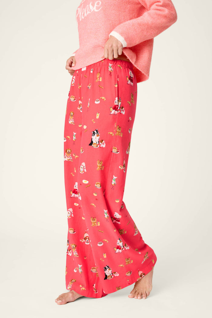 A person wearing a pink sweater and P.J. Salvage Dogs Who Brunch Flannel Pajama Pants, featuring playful prints of dogs and brunch foods, stands barefoot against a plain light background.