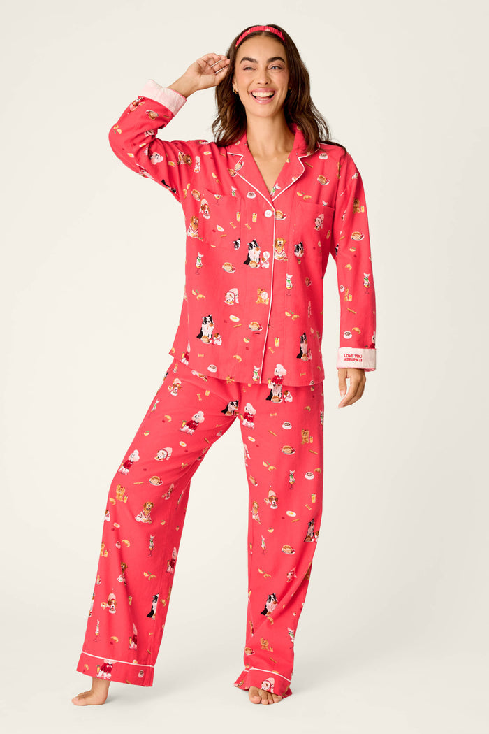 A woman stands barefoot against a light beige background, playfully touching her hair and smiling as she models the P.J. Salvage Dogs Who Brunch Print 3-Piece Long Sleeve Flannel PJ Set. She wears the cozy red cotton pajamas adorned with whimsical dog-themed prints and completes her look with a matching red headband, radiating a relaxed, dog-lover style.