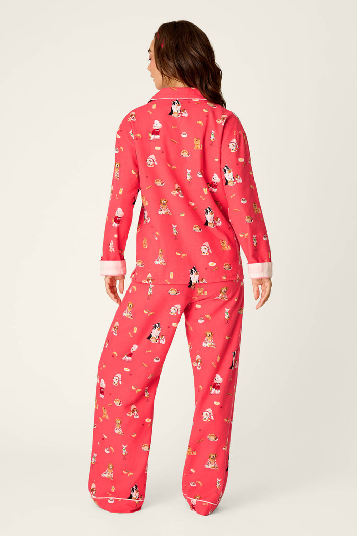 A woman stands facing away, wearing the P.J. Salvage Dogs Who Brunch Print 3-Piece Long Sleeve Flannel PJ Set, featuring a red cotton flannel material with white cuffs and a playful pattern of various cartoon dogs and bones. The background is plain and light-colored.