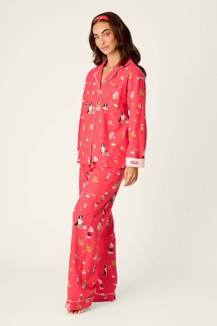 A woman stands and smiles in front of a plain light background, wearing the P.J. Salvage Dogs Who Brunch Print 3-Piece Long Sleeve Flannel PJ Set — a bright pink cotton flannel ensemble featuring charming cartoon dog prints, matching pants, and a coordinating pink headband.