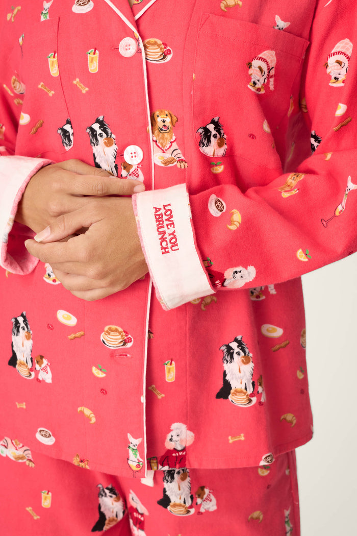 A person wears the P.J. Salvage Dogs Who Brunch Print 3-Piece Long Sleeve Flannel PJ Set, featuring bright pink cotton flannel with playful breakfast food and dog patterns; their hands are folded, and the sleeve reveals a white cuff accented with red “LOVE YOU ABUNCH” lettering.