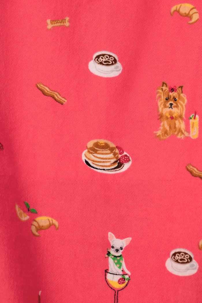 A close-up of a pink fabric with a repeating pattern of small illustrated food and animal motifs, including pancakes with strawberries, croissants, a dog, a cat in a glass, and coffee cups.