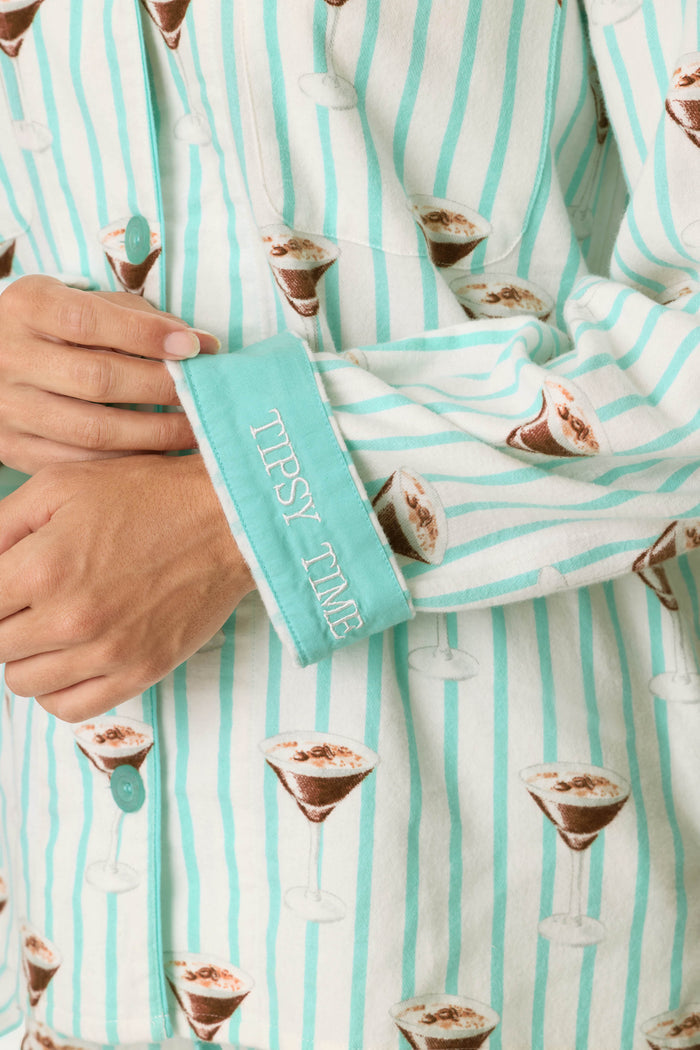 A person wearing the P.J. Salvage Espresso Martini Print 3-Piece Long Sleeve Flannel PJ Set, featuring blue stripes and martini glass prints, with the cuff flipped to reveal embroidered text reading “TIPSY TIME.”.