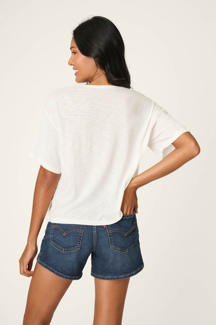A woman with long dark hair stands smiling with her back to the camera, wearing the P.J. Salvage Feelin’ Prickly Spicy Chili Graphic T-Shirt and blue denim shorts, her hand on her hip against a light background.