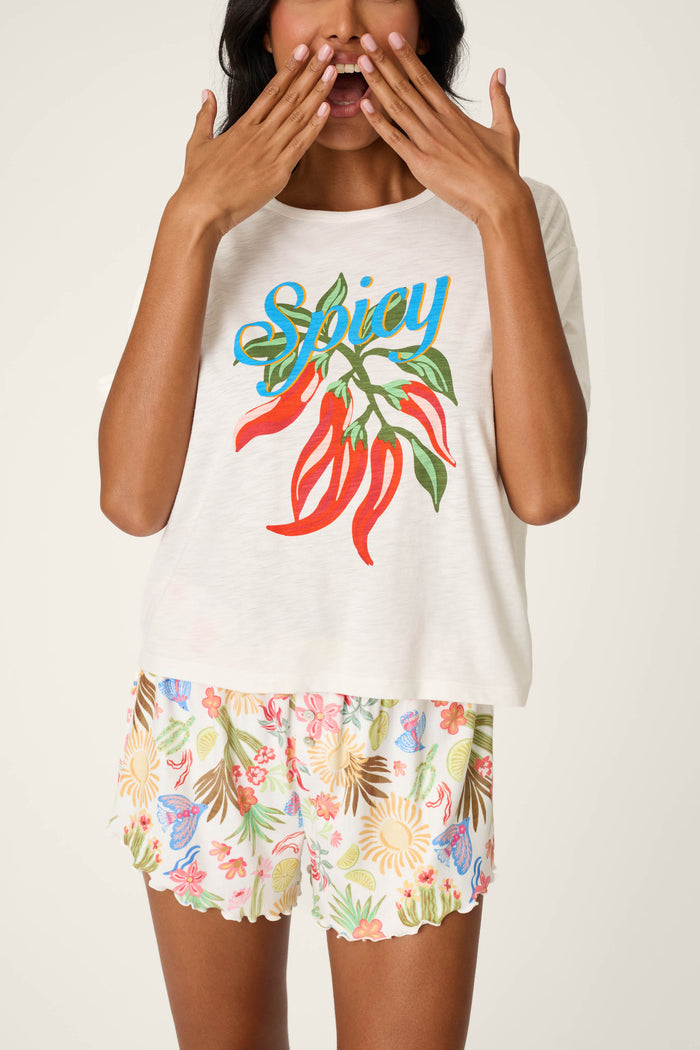 A woman playfully poses against a light background, wearing the P.J. Salvage Feelin’ Prickly Spicy Chili Graphic T-Shirt and floral shorts. She holds her hands by her mouth, showcasing the shirt's bold chili graphic.
