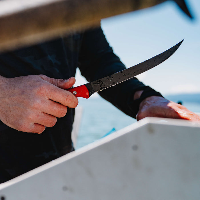 A person holding a red-handled Flathead fillet knife while preparing fish on a boat by the water.