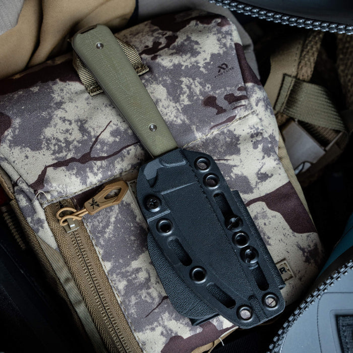 Olive green FLATTAIL knife sheathed in a black Kydex holder, resting on a camo-patterned bag. Ideal for outdoor use.