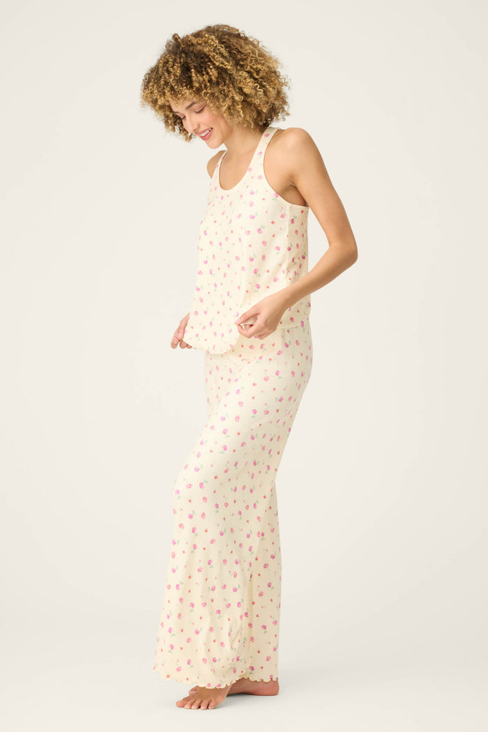 A person stands barefoot on a light background, smiling and gently holding the hem of their For the Mamas Butter Blooms Long PJ Set—a sleeveless, cream-colored floral pajama set by P.J. Salvage.