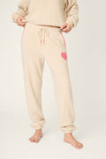 A person models the P.J. Salvage For the Mamas Sweetheart Banded Pants—plush, cream-colored fleece joggers with a pink heart on the left thigh and red-and-white checked pocket accents, shown barefoot from the waist down.