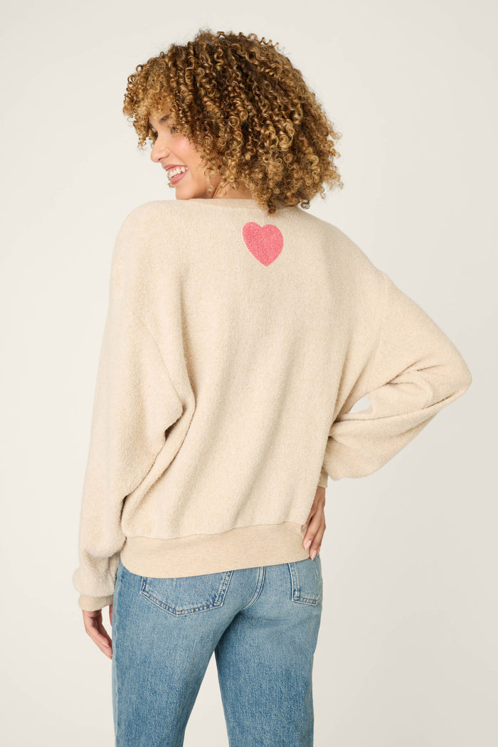A woman with curly hair models the P.J. Salvage For the Mamas Sweetheart Mama Sweatshirt in light oatmeal, featuring a small pink heart on the upper back. She pairs it with light blue jeans. Made from cloud brushed terry for comfort.