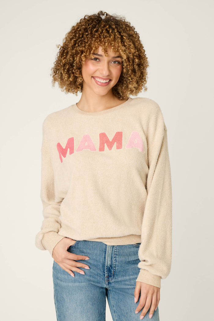 A person with curly hair smiles at the camera, wearing the P.J. Salvage For the Mamas Sweetheart Mama Sweatshirt in light oatmeal, paired with blue jeans, and stands against a plain light background.
