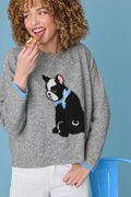 A smiling person with curly hair enjoys a pastry while wearing the P.J. Salvage French Bulldog Snuggle Sweater, which features a black and white dog design and light blue trim, standing against a blue background.