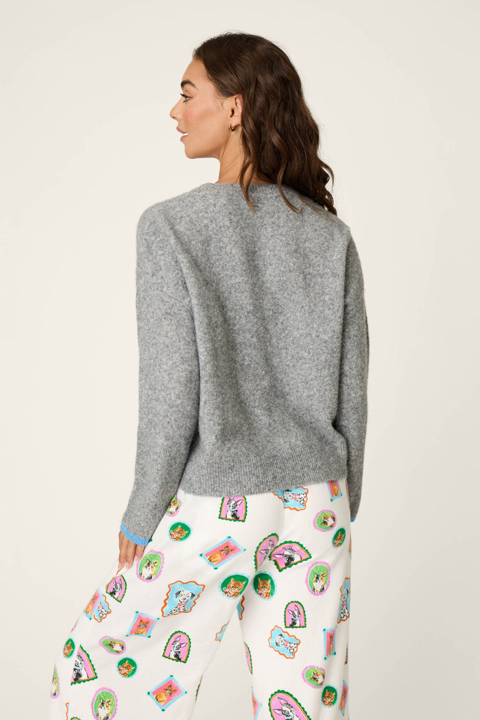 A woman faces away, wearing the P.J. Salvage French Bulldog Snuggle Sweater in gray paired with white pants adorned with colorful cartoon faces and shapes. The background is plain and light-colored.
