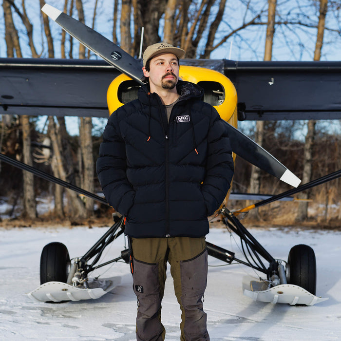 Man wearing a black Grinnell Puffy Zip Jacket stands in front of a yellow aircraft in a snowy landscape.
