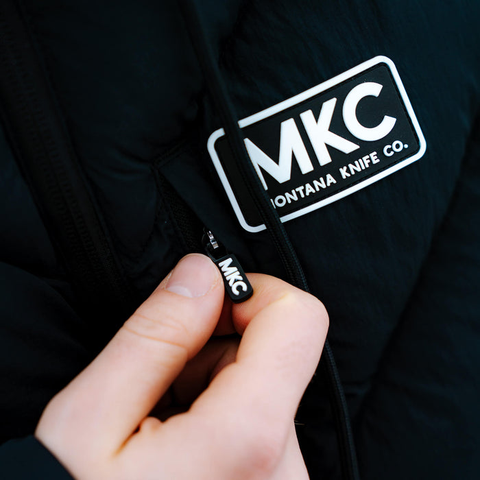 Close-up of a person zipping the black Grinnell Puffy Zip Jacket featuring the Montana Knife Company logo.