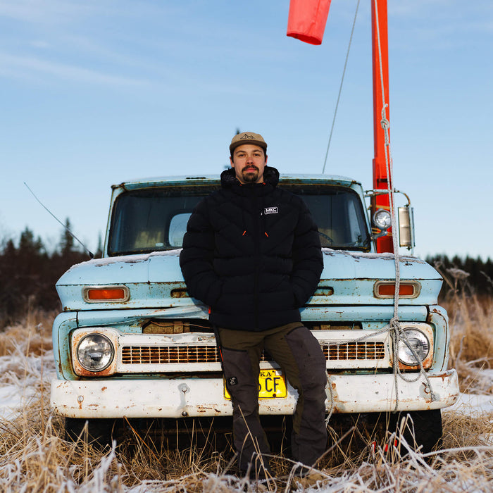 A man wearing a black Grinnell Puffy Zip Jacket stands on the hood of an old truck in a snowy landscape.