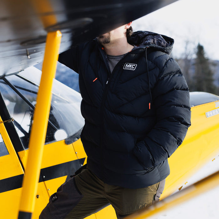 Montana Knife Company black Grinnell Puffy Zip Jacket worn by a person near a yellow airplane in a snowy outdoor setting.