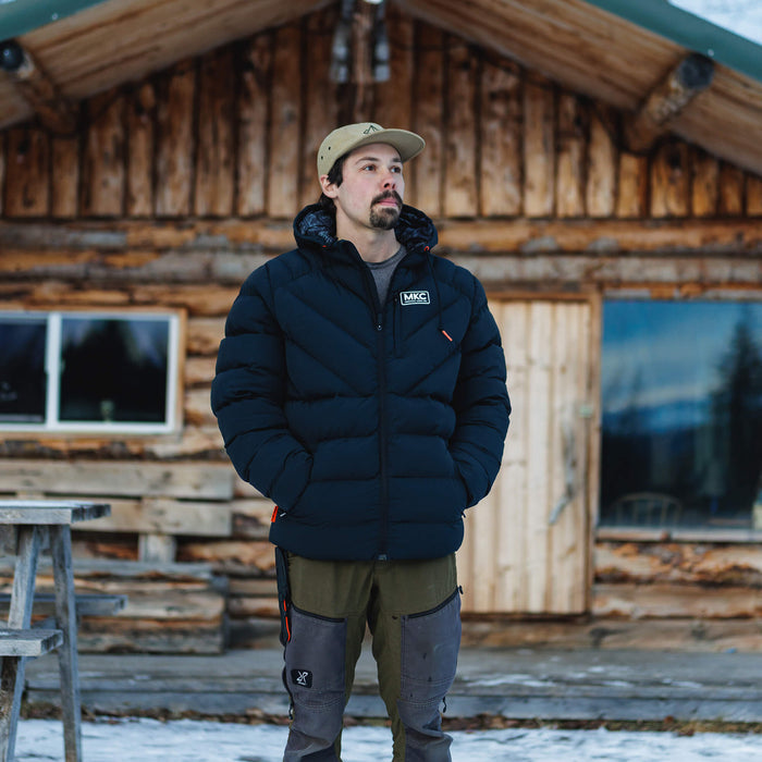 Man wearing a black Grinnell Puffy Zip Jacket outside a rustic wooden cabin, showcasing warmth and outdoor style.
