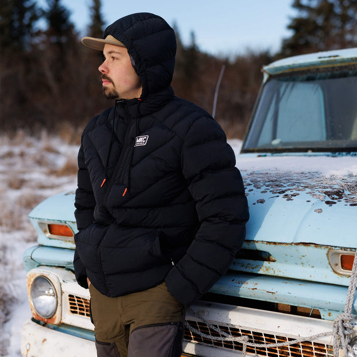 A man wearing a black Grinnell Puffy Zip Jacket stands beside a vintage truck in a snowy landscape.