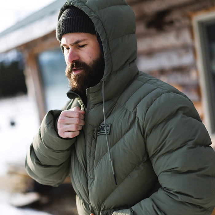 Man wearing an olive green Grinnell Puffy Zip Jacket, adjusting the zipper outdoors in a snowy setting.