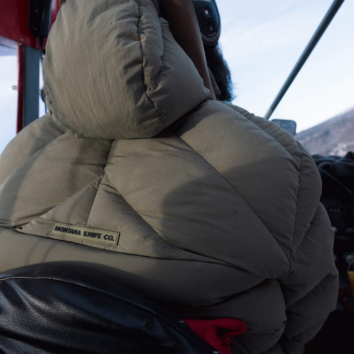 Olive Grinnell Puffy Zip Jacket from Montana Knife Company, featuring a hood and logo, worn in an outdoor setting.