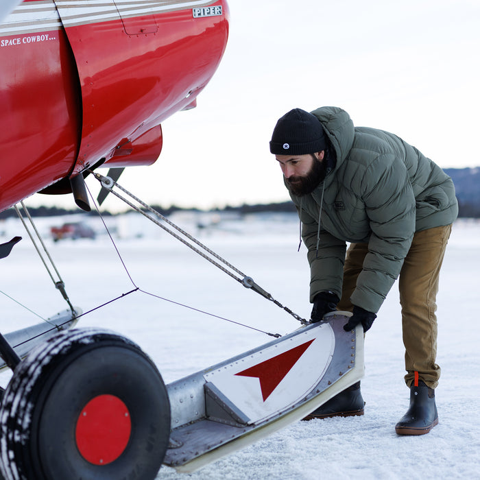 Man wearing an olive GRINNELL PUFFY ZIP JACKET loading gear onto a red airplane on a snowy runway.