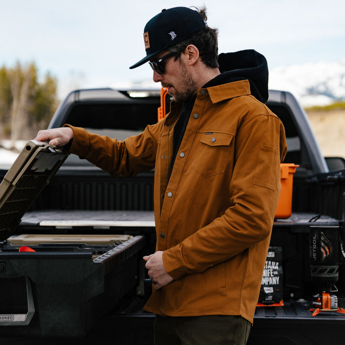 Man wearing a coyote-colored Hamilton shacket, inspecting gear in a truck bed with mountains in the background.