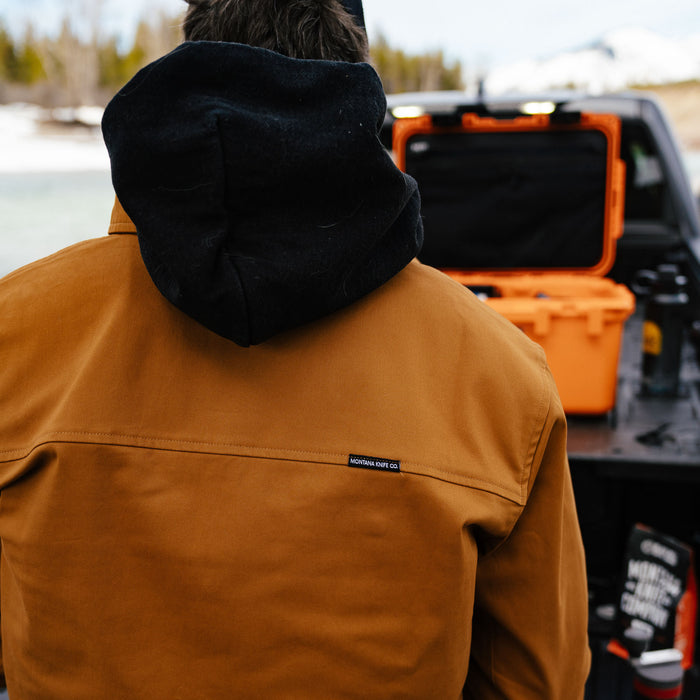 A person wearing a coyote-colored Hamilton shacket stands by a truck, with a scenic outdoor backdrop.