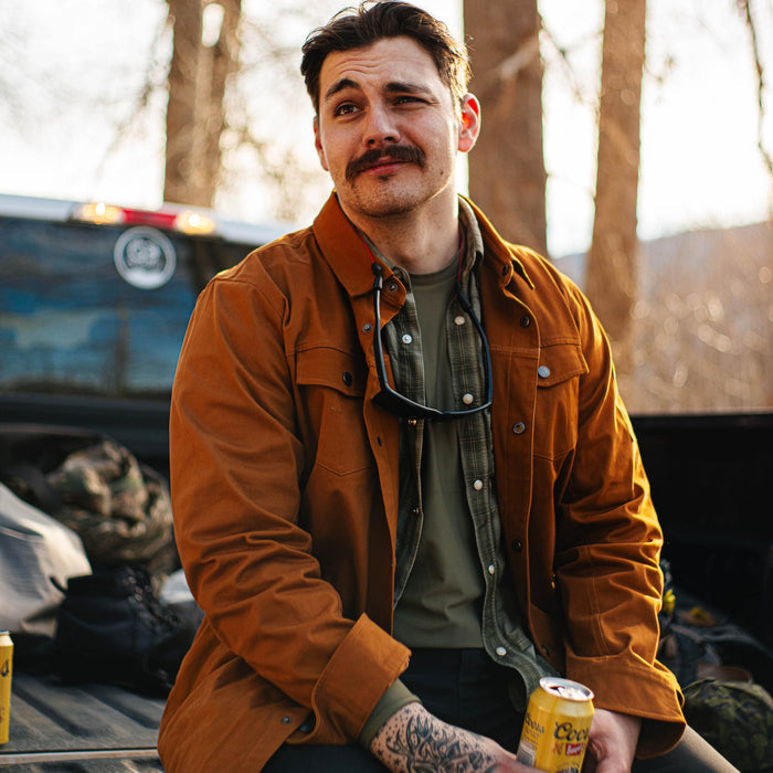 A man wearing a coyote-colored Hamilton shacket sits on a truck bed, holding a drink, with trees in the background.