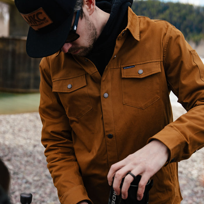 A man wearing a coyote-colored Hamilton shacket, adjusting equipment outdoors near a river.