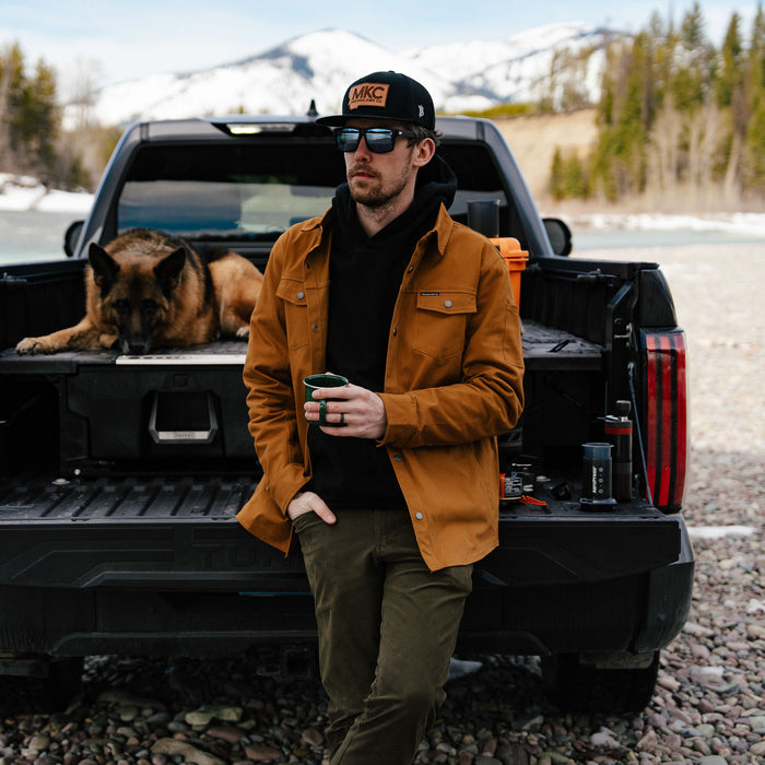 Man wearing a coyote-colored Hamilton shacket stands by a truck, holding a cup, with a dog resting in the truck bed.