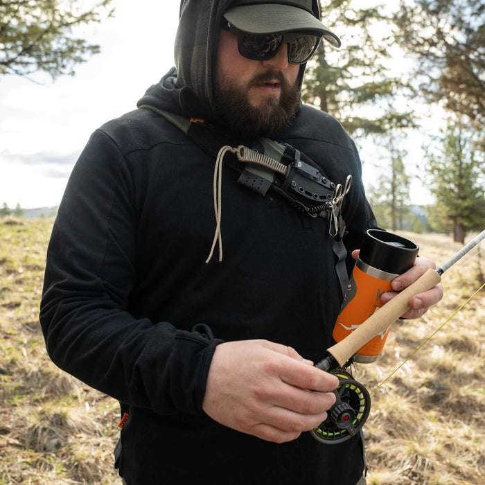 A man wearing a black Highline half-zip hoodie adjusts a fishing reel while holding a coffee cup outdoors.