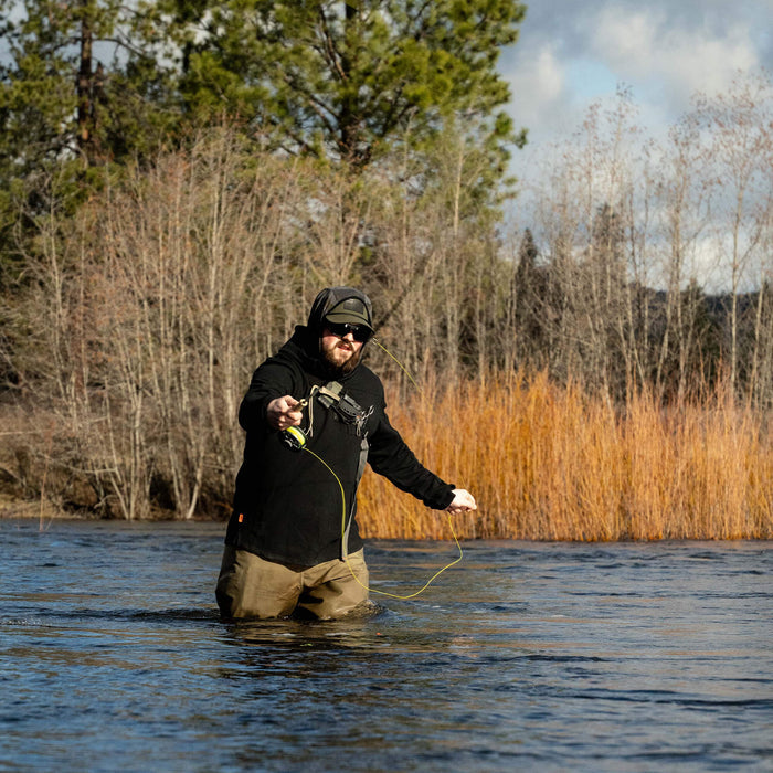 Man wearing a black Highline half-zip hoodie, wading in a river while fly fishing amidst trees and tall grass.