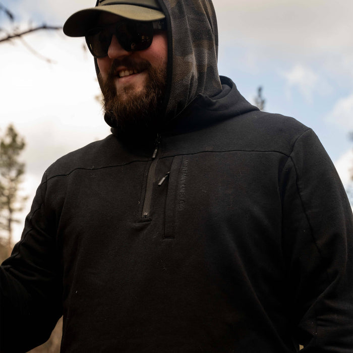 Man wearing a black Highline half-zip hoodie with a hood, outdoors in a forested area, smiling and enjoying nature.