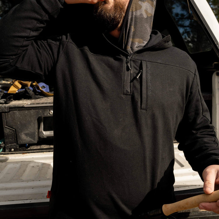 Highline half-zip hoodie in black, featuring a camo-lined hood, worn by a man outdoors. Ideal for work and outdoor activities.