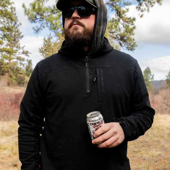 Highline half-zip hoodie in black worn by a man outdoors, holding a can, surrounded by trees and grass.