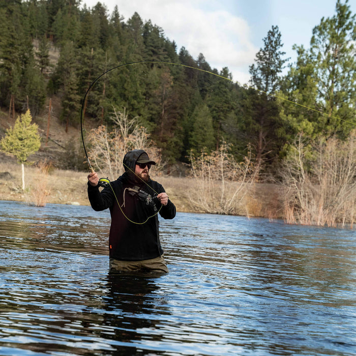 Man fishing in a river while wearing a black Highline half-zip hoodie, surrounded by trees and mountains.