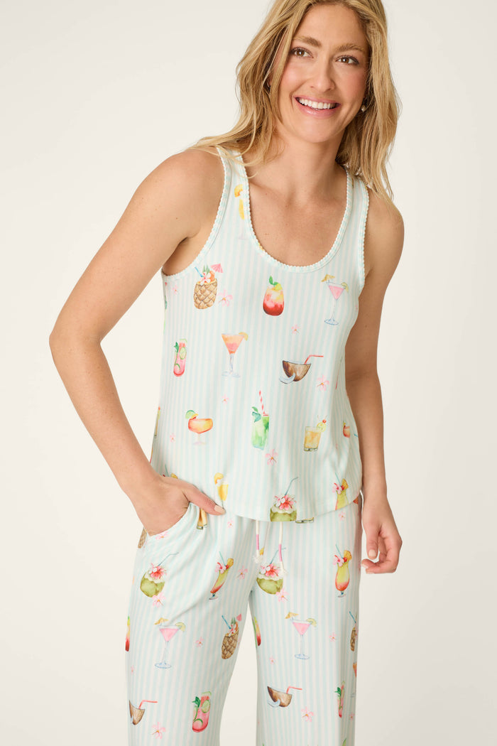 A woman smiles with one hand in her pocket, wearing the P.J. Salvage It's Five O'clock Somewhere Cocktail Print Tank Top—a sleeveless, light-colored pajama top featuring colorful tropical drinks and fruits.