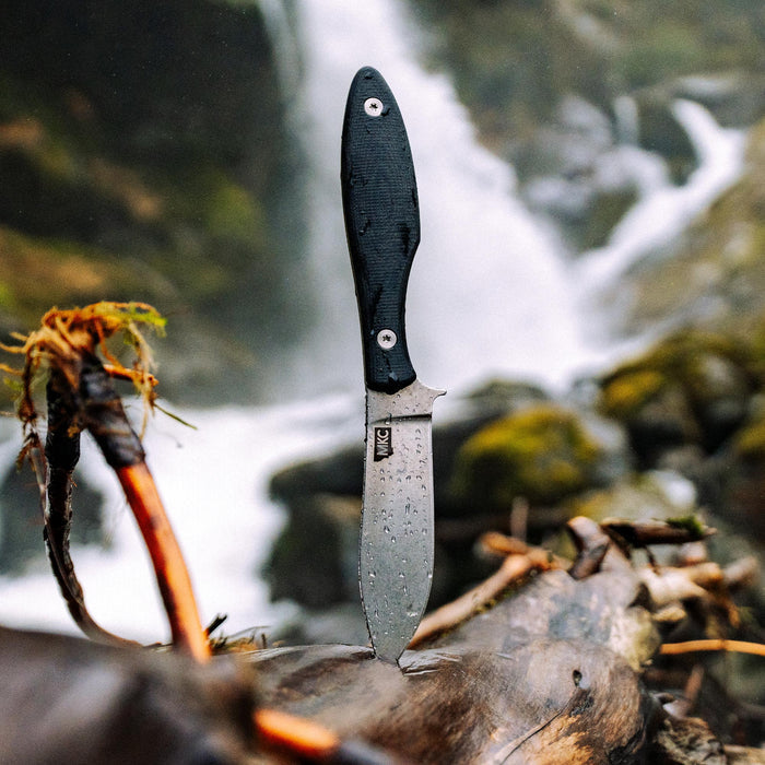 Black JACKSTONE knife with a textured handle, displayed on a log near a waterfall, showcasing its rugged design.