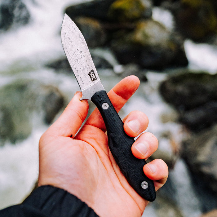 Hand holding the JACKSTONE Black knife with a textured handle, set against a natural rocky background.