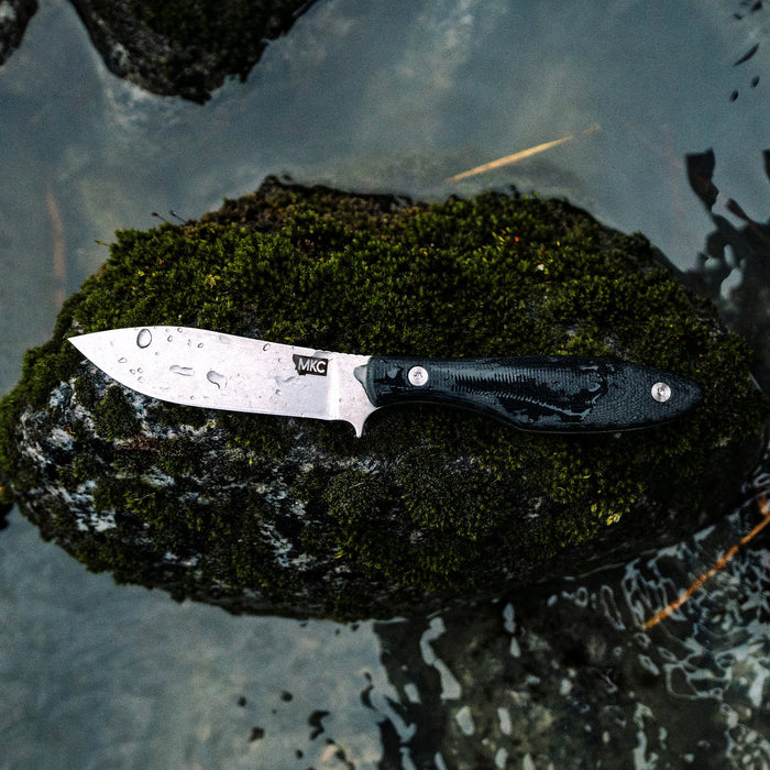 Black JACKSTONE knife resting on a mossy rock near water, showcasing its sleek blade and textured handle.
